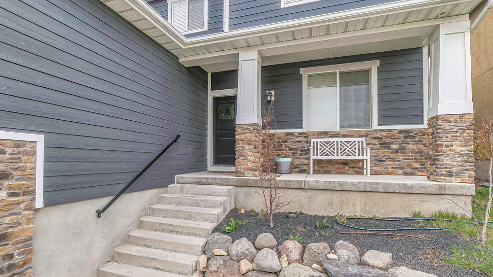 Pano Stairs going up the porch of home with gray front door and window at the facade. White bench, windows, wooden siding, and brick wall can also be seen at the exterior.